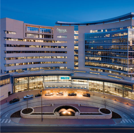 A large, modern hospital building with many windows is lit up at dusk. Several cars are parked in front, and a curved driveway and landscaped area are visible near the entrance.