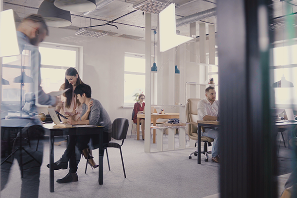 Modern open-plan office with people working at desks, collaborating in small groups. Large windows provide natural light, and the workspace features contemporary furniture and decor. A person is walking past in the foreground.