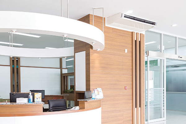 A modern, clean reception area with a curved white and wood desk, office chairs, computer monitors, and a glass entrance door. The space is brightly lit with minimalist decor and a mounted air conditioner.