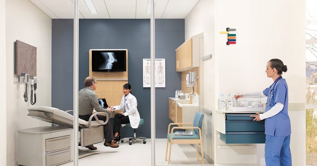 A doctor talks with a patient seated on an exam table in a medical office while a nurse in scrubs pushes a cart nearby. Medical equipment and an X-ray image are visible in the room.