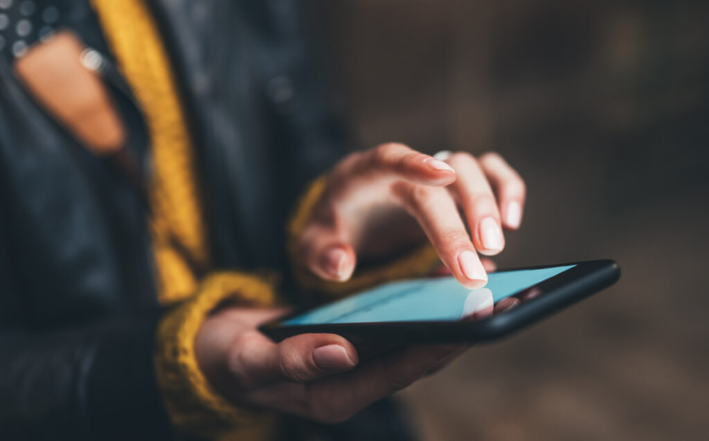 Close-up of a person’s hands using a smartphone. The person is wearing a dark jacket and a yellow sweater, and is touching the screen with their index finger. The background is blurred.