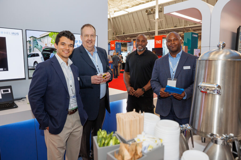 Four men in business attire stand and smile at a conference or trade show booth. In the foreground, there are cups, stir sticks, and a large beverage dispenser on a table. The background shows other booths and displays.