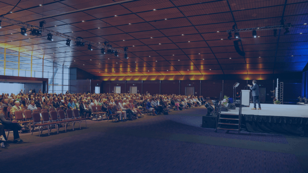 A large audience sits in rows facing a stage where a person is speaking at a podium in a spacious, modern conference hall with high ceilings and warm lighting.
