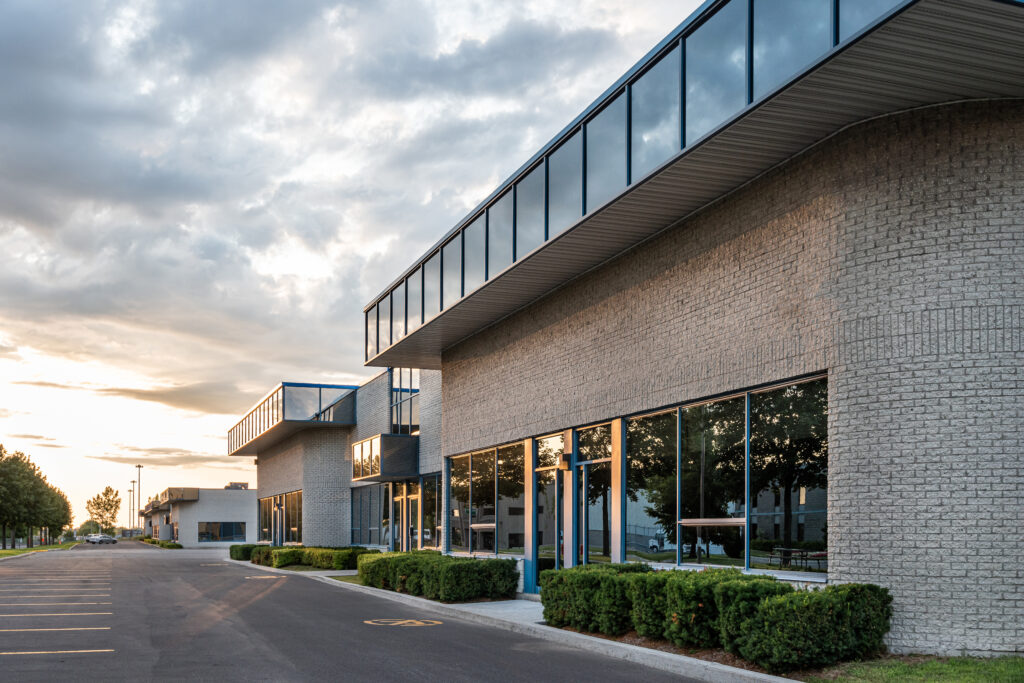 Modern commercial office buildings with large windows and light brick exteriors, reflecting trees and sky at sunset. Manicured bushes line the walkway, and the area appears quiet and empty.