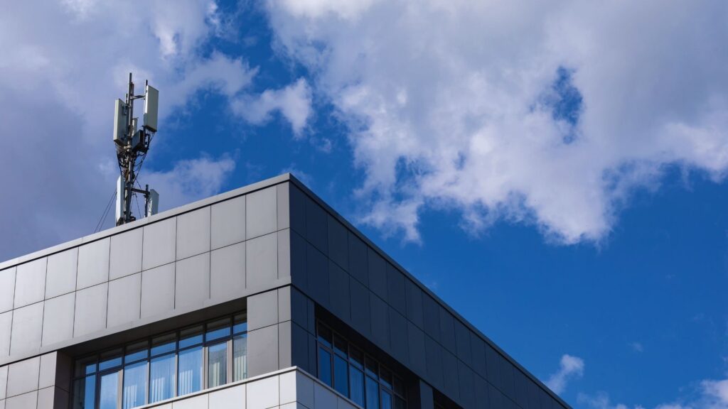A gray modern building with a cellular antenna on its roof, set against a bright blue sky with scattered white clouds.