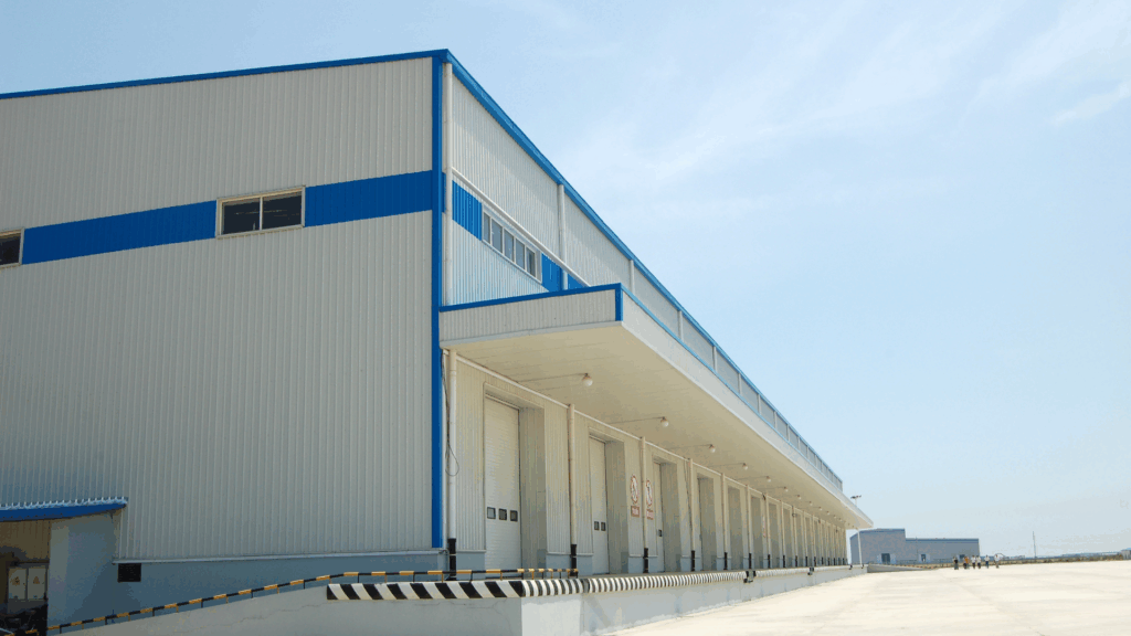 A large industrial warehouse with white and blue exterior, multiple loading dock doors, and a wide concrete pavement under a clear blue sky.