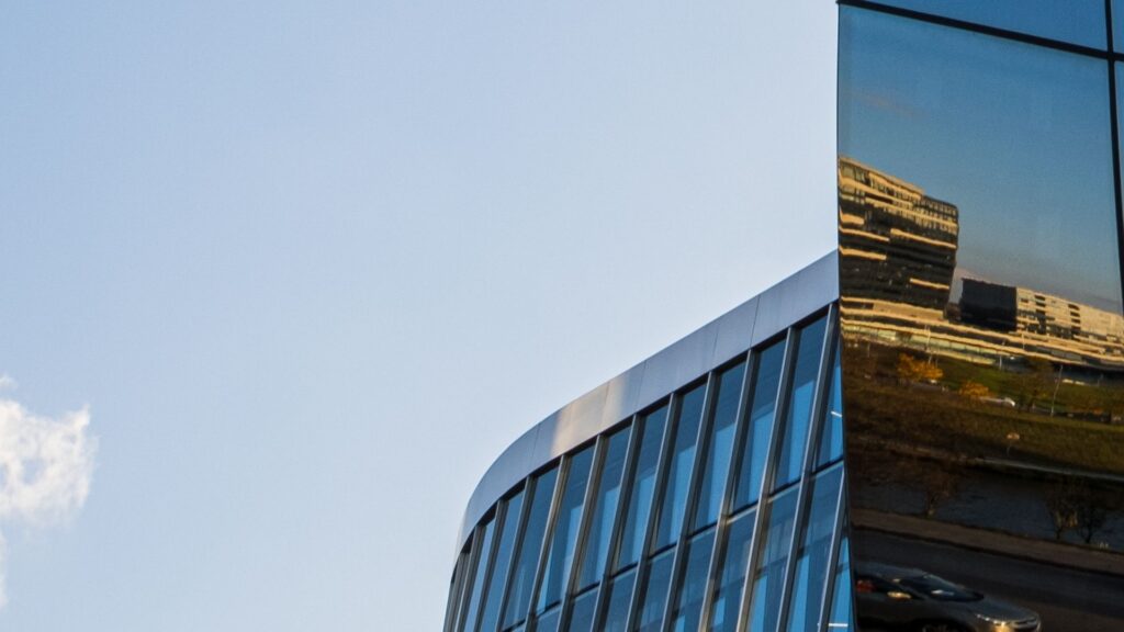A modern glass building with curved edges reflects another building and part of the cityscape in its windows. The sky is clear and blue with a small cloud on the left.