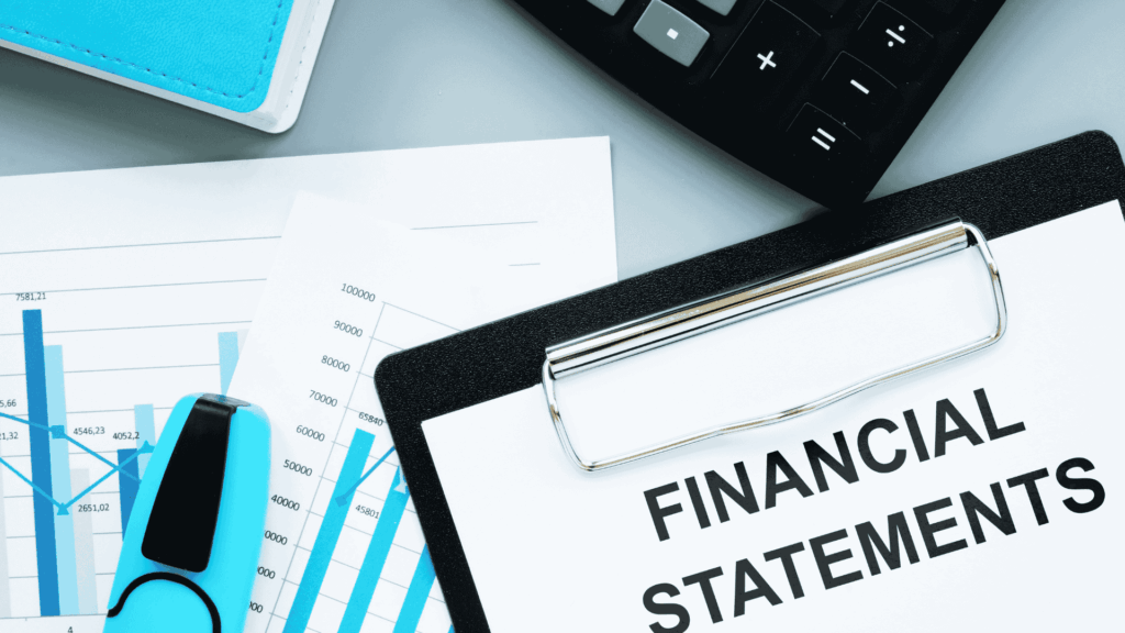 A clipboard with a paper titled FINANCIAL STATEMENTS lies on a desk next to a calculator, a blue binder, a highlighter, and printed charts with blue bars and numbers.