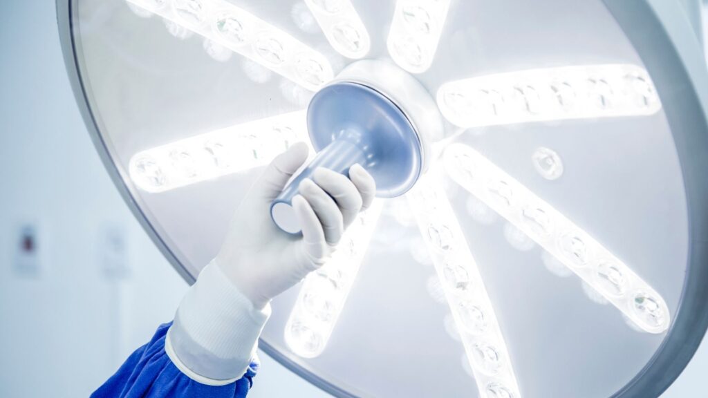 A gloved hand adjusts a large, circular surgical light with multiple bright LED bulbs in a medical or hospital setting.