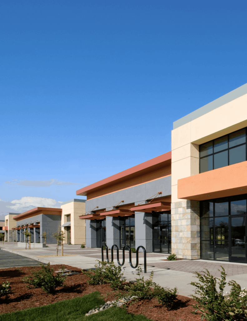 A row of modern, empty commercial storefronts with large windows, orange accents, and a bike rack in front. The landscaping is neatly maintained, and the sky is clear and blue.