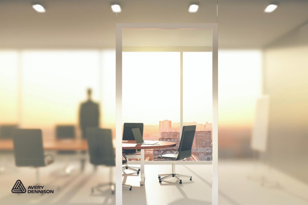 A modern office with large windows showing a city view at sunset. Empty chairs surround a conference table with a laptop. A blurred figure stands in the background. Avery Dennison logo is in the lower left corner.