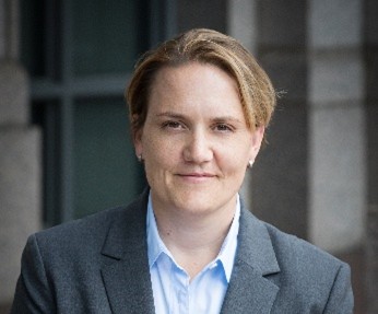 A person with short light brown hair, wearing a gray blazer and light blue shirt, stands in front of a stone building and looks at the camera with a slight smile.