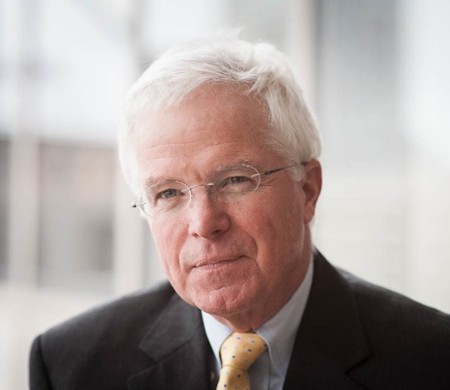 An older man with short white hair and glasses, wearing a dark suit, white shirt, and yellow polka dot tie, looks thoughtfully to the side in a softly lit indoor setting.
