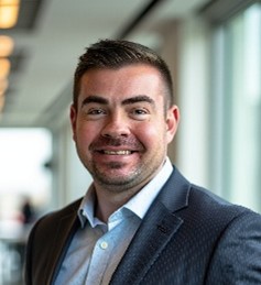 A man with short dark hair and a trimmed beard is smiling, wearing a dark blazer and light blue shirt, standing indoors next to large windows with blurred lights in the background.