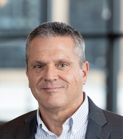 A middle-aged man with short gray hair, wearing a gray suit jacket and a white checkered shirt, smiles slightly while standing indoors in front of large windows.