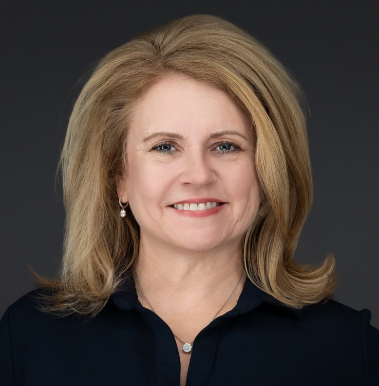 A woman with shoulder-length blonde hair, wearing a black blouse, a necklace, and small earrings, smiles at the camera against a plain dark background.
