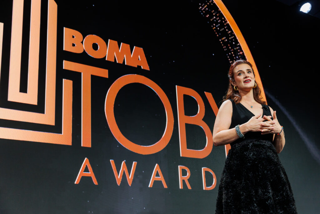 A woman in a black dress speaks onstage at the BOMA TOBY Awards, standing in front of a large illuminated event logo.