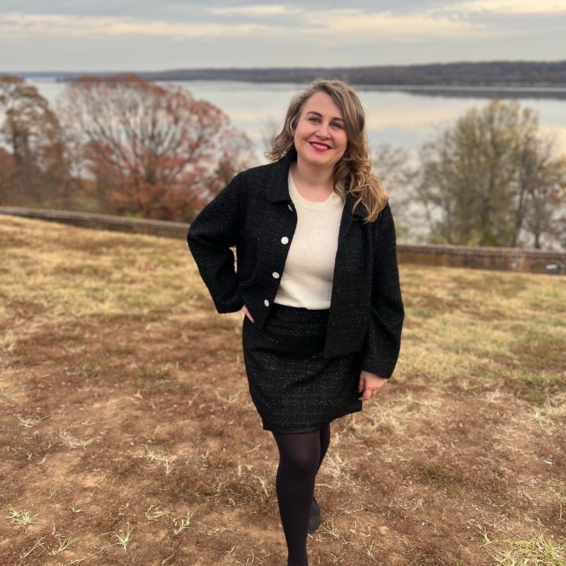 A woman in a black jacket and skirt, white top, and black tights stands smiling outdoors on a grassy area with a lake and trees in the background.