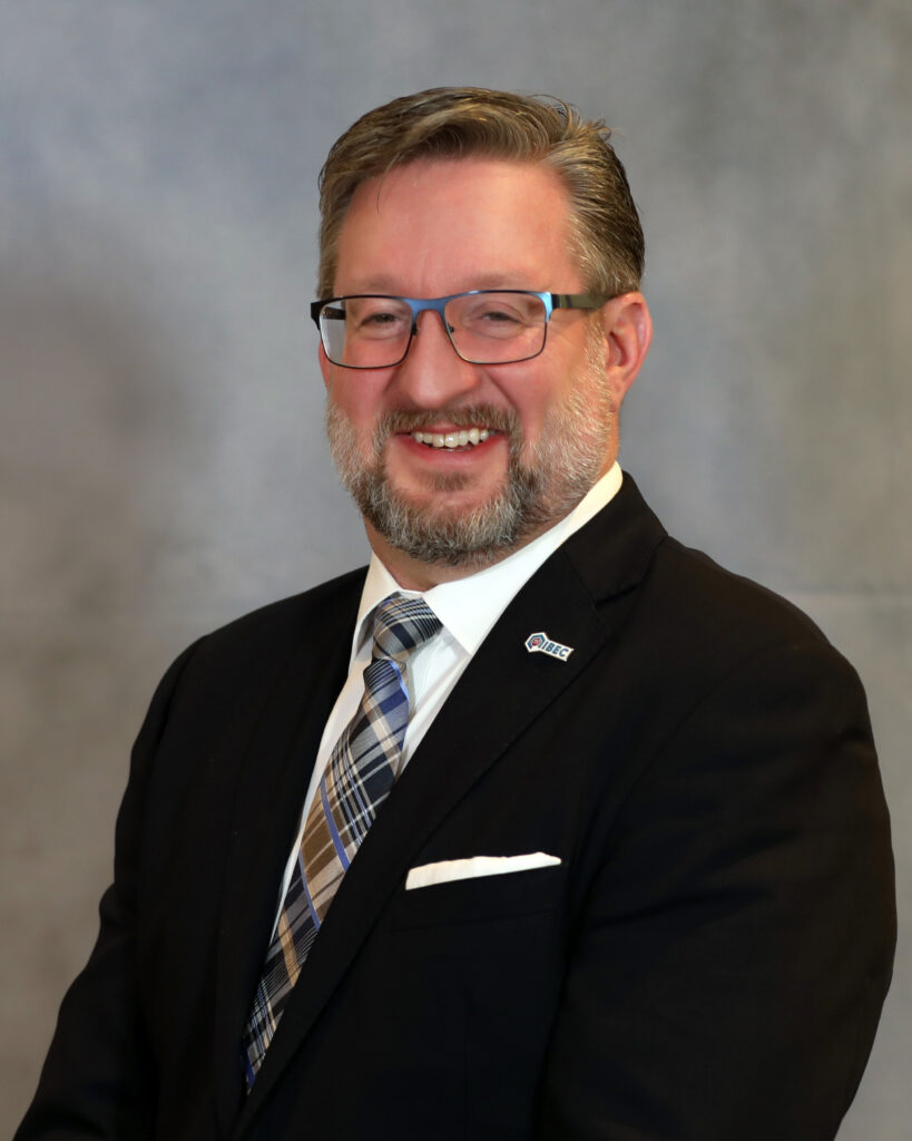 A man with glasses and a beard, wearing a dark suit, plaid tie, white shirt, and pocket square, smiles in front of a neutral gray background.