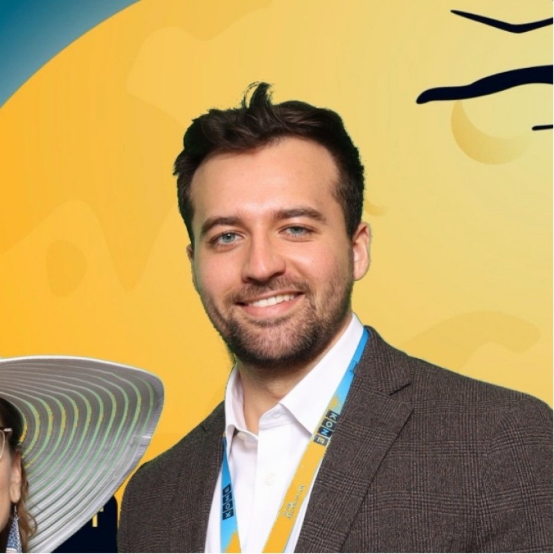 A man in a brown blazer and light blue shirt smiles in front of a yellow abstract background. He wears a conference lanyard; part of a large hat is visible on the left.