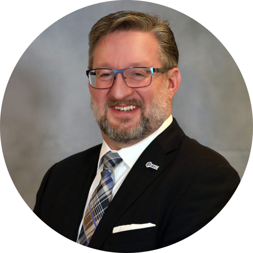 A smiling man with a beard and glasses, wearing a dark suit, white shirt, and plaid tie with an IIBEC pin on his lapel.